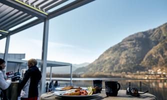 A table with a breakfast plate and two cups of coffee is in the foreground. In the background, mountains and a clear sky can be seen.