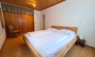 A simple bedroom with a double bed and wooden floor. The walls are light and there is a chair as well as a wardrobe.