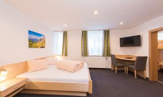 A modern room with a double bed and a small desk.  
Large windows with curtains provide plenty of daylight.