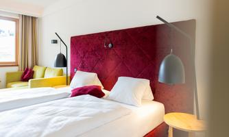 A modern hotel room with two single beds and a red headboard. The decor is stylish with a cozy green seating area and elegant lamps.