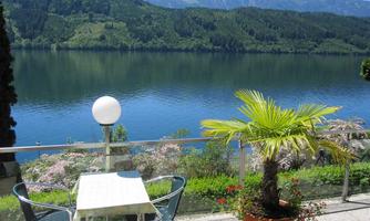 A beautiful view of a tranquil lake, surrounded by green hills. On the terrace, there is a table and chairs, as well as a palm in a pot.