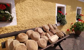 A bench with several artistically crafted woven baskets stands in front of a light yellow wall. Flowers bloom in the windows and beside it.