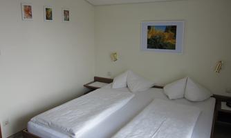 A simple bedroom with two single beds and decorative cushions. Some pictures hang on the wall, and there is soft lighting.