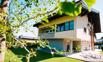 A modern house with large windows and a well-maintained green area. In the foreground, fresh green leaves can be seen.