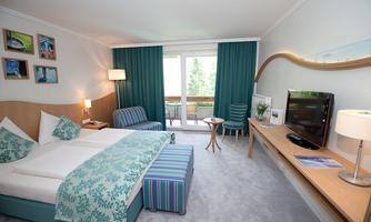 A bright hotel room with a double bed and colorful decorations. There is a balcony overlooking nature and modern furniture.