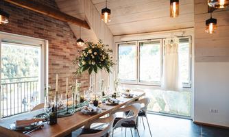 A stylish dining area with a long wooden table and elegant chairs. Large windows offer a view of nature and the table is set festively.