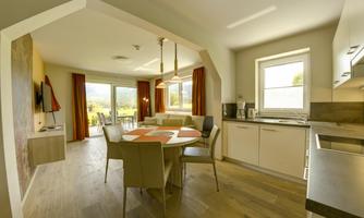 A modern kitchen with a dining area and an open living space. Large windows offer a view of the outdoor terrace and the surroundings.