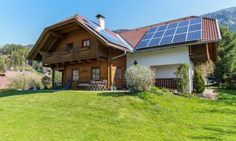 A beautiful house with a garden and solar panels on the roof. The surroundings are green and surrounded by mountains.