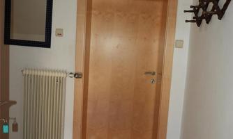 A hallway with a wooden door and a radiator. On the wall hangs a mirror and a wall hook, and a clock is mounted on the wall.