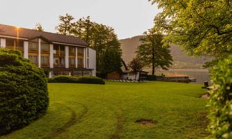 A quiet property by the water with green grass and trees. The sun rays shine on the building in the background.