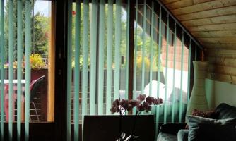 A cozy living room with a large window front and modern vertical blinds. In the background, plants and a terrace are visible.