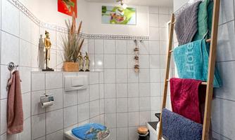 A modern bathroom with white tiles and a sink. Towels in various colors hang on a wooden shelf and there are decorative pictures on the wall.