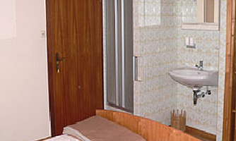 A simple bedroom with a bed and light blankets. In the background, a bathroom with a sink and shower is visible.