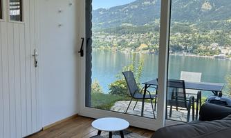 A cozy living room with a view of a tranquil lake and surrounding mountains. Outside, there are chairs and a table on a terrace.