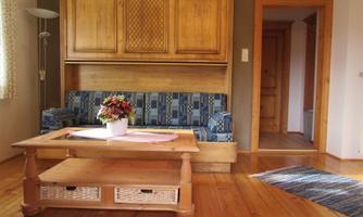 A cozy living room with a blue sofa and a wooden table. The room has wooden floors and simple wall decoration.