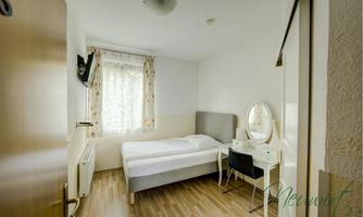 A simple bedroom with a bed, a table, and a mirror. There is a window with curtains and a television on the wall.
