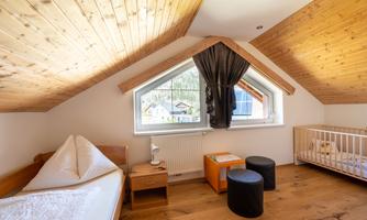 A modern room with a wooden ceiling and a window that lets in lots of daylight. The room contains a bed, a children's playpen, and two seating areas.