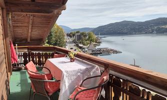 A beautiful balcony with a table and chairs, overlooking a tranquil lake and the surrounding mountains. In the background, boats and a small town are visible.
