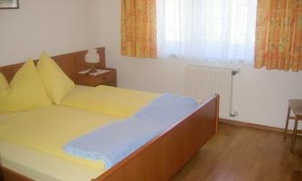 A simple bedroom with two beds, yellow pillows, and a blue blanket. The room has large windows with curtains in warm colors.