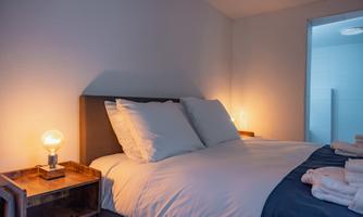 A cozy bedroom with a large bed and two pillows. Next to the bed is a lamp and the room has a warm, welcoming atmosphere.