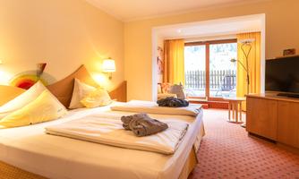 A cozy bedroom with two beds, soft pillows, and a television. Large windows bring in plenty of light and provide access to a balcony.