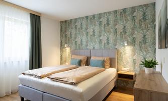 A modern bedroom with a double bed and elegant leaf-patterned wallpaper. The room is bright and welcoming, with natural light and minimalist decor.