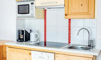 A modern kitchen with wooden furniture and white ceramic tiles. The equipment includes a stove, a sink, a microwave, and a kettle.