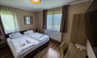 A cozy hotel room with two beds, a desk, and a television. Large windows provide plenty of daylight.
