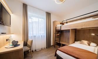 A modern room with wooden furniture and a bunk bed. There is a desk, a television, and plenty of natural light coming through the window.