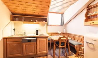 A cozy kitchen with wooden cabinets and a dining table. The bright atmosphere is emphasized by the sloping roof and the windows.