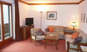 A cozy living room with a sofa, armchairs, and a table. The decor is in warm colors and there is a television as well as a window with access to the balcony.