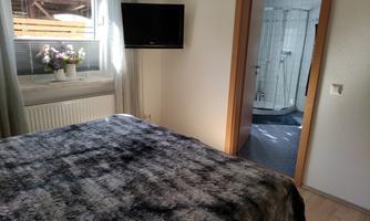 A cozy bedroom with a window, a television, and a double bed. In the background, you can see a modern bathroom.
