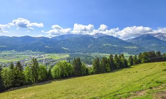 A picturesque landscape with green meadows and high mountains in the background. The sky is clear and blue, with some white clouds.