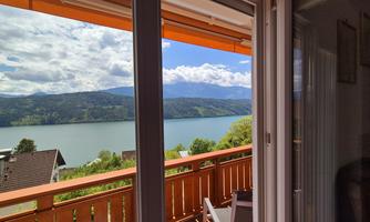 A view from a balcony of a tranquil lake and surrounding mountains. The sky is partly cloudy with some rays of sunshine.