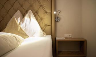 A cozy bedroom with a padded headboard and two pillows. Next to the bed is a wooden shelf with a lamp and an outlet.