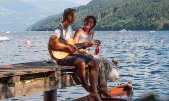 A couple is sitting on a dock by the water and playing guitar. The atmosphere is relaxed and romantic, surrounded by nature and mountains.