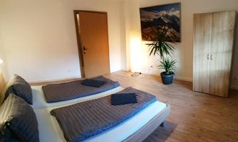 A cozy bedroom with two beds and modern decorations. The room is bright and friendly, with a window and a plant arrangement.