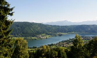 A picturesque landscape with a clear lake and green hills. In the distance, mountains and a small village can be seen.
