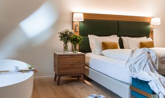 A cozy bedroom with a large bed and wooden nightstands. The room features a bathtub and fresh flowers on the table.