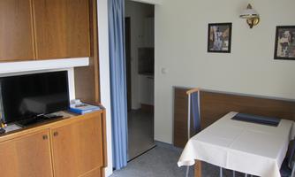 A cozy living area with a television, a wooden table, and pictures on the wall. The room has bright walls and access to another area.