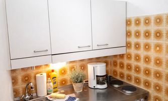 A small kitchen with modern, white cabinets and a decorative wall tile. On the countertop, there is a coffee machine and cleaning supplies.