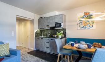 A modern kitchen with gray furniture and a dining table. The space is cozily decorated and features a hanging light circle.