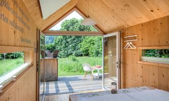 A bright wooden house with large windows and a terrace that leads into nature. Interior with a cozy bed and simple furnishings.