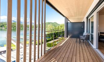 A modern balcony with a wooden terrace overlooking a peaceful lake. In the background, green hills and blue sky are visible.