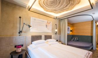 A modern hotel room with a double bed and a cozy seating area. The walls are painted in soft colors and adorned with appealing decorations.