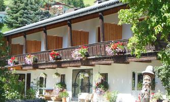 A charming building with a wooden balcony adorned with colorful flowers. In the foreground, there are decorative chairs and a large wood carving.