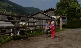 A child is standing next to a woman and looking at a goat. In the background, farms and green hills are visible.