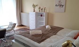 A cozy bedroom with a large bed, fresh towels, and a modern armchair. There is a desk and a bright wardrobe in the background.