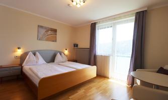 A bright hotel room with two single beds and a large window. The floor is made of wood, and there is a small table and chairs in the room.