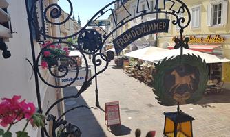 A decorative sign with the inscription "Guest Room" hangs over a small street. In the background, cafes and flowers can be seen, creating an inviting atmosphere.